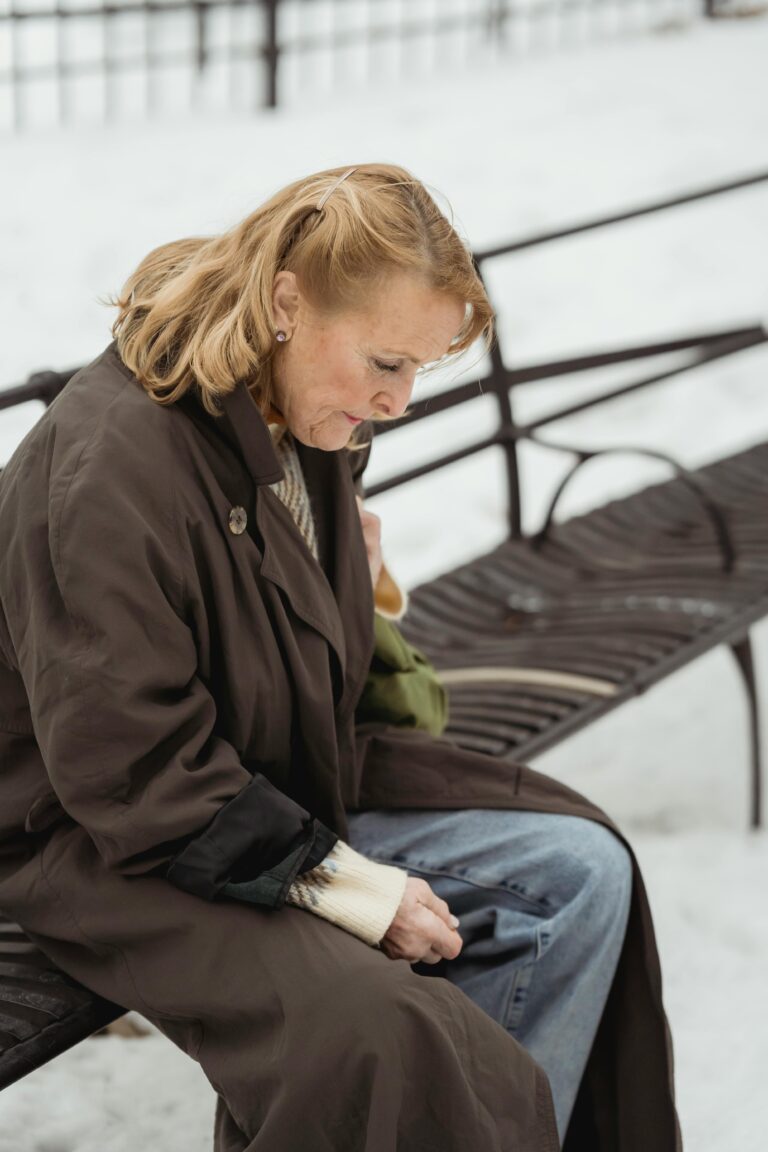 Side view of hopeless senior female in outerwear looking down while sitting on bench in winter city