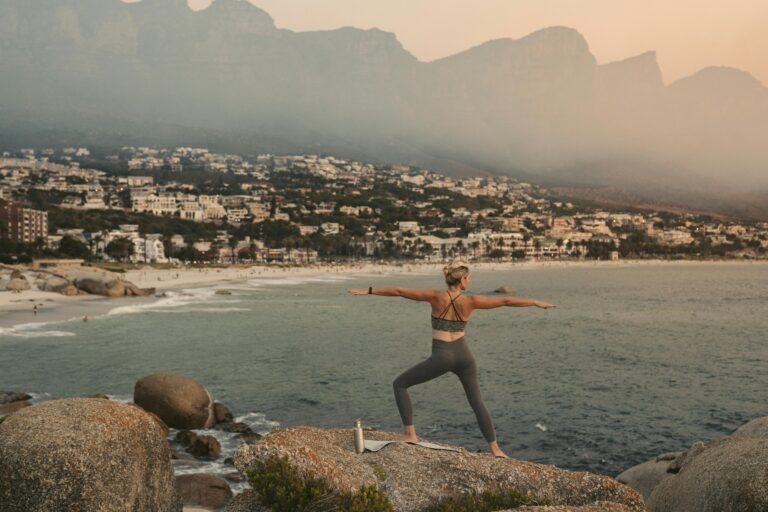 Woman practicing yoga warrior pose with scenic view of Cape Town coastline at sunset, promoting wellness.